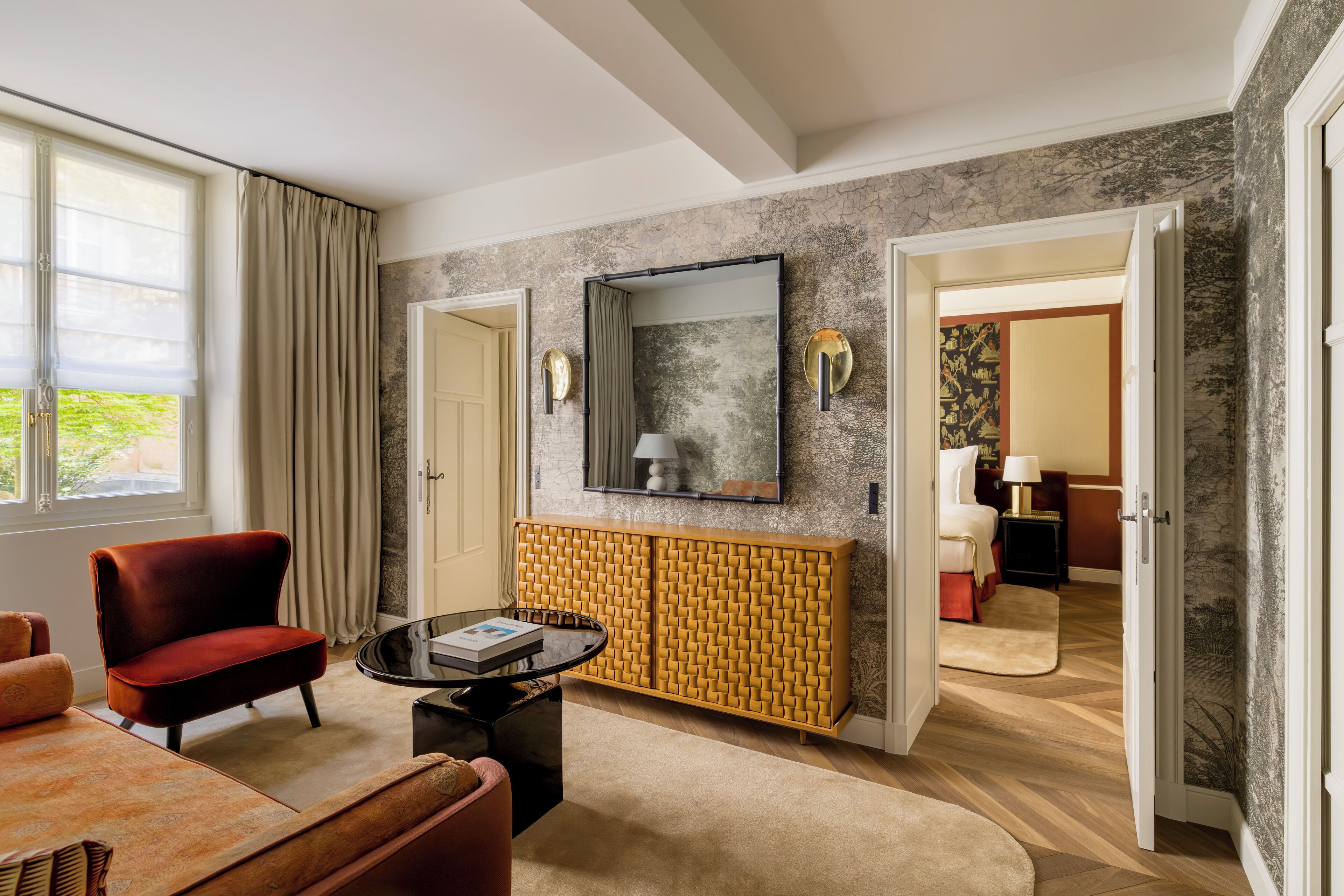 A hotel room with a modern design, featuring a red chair, a round coffee table, a yellow patterned console, and a large mirror on the wall.