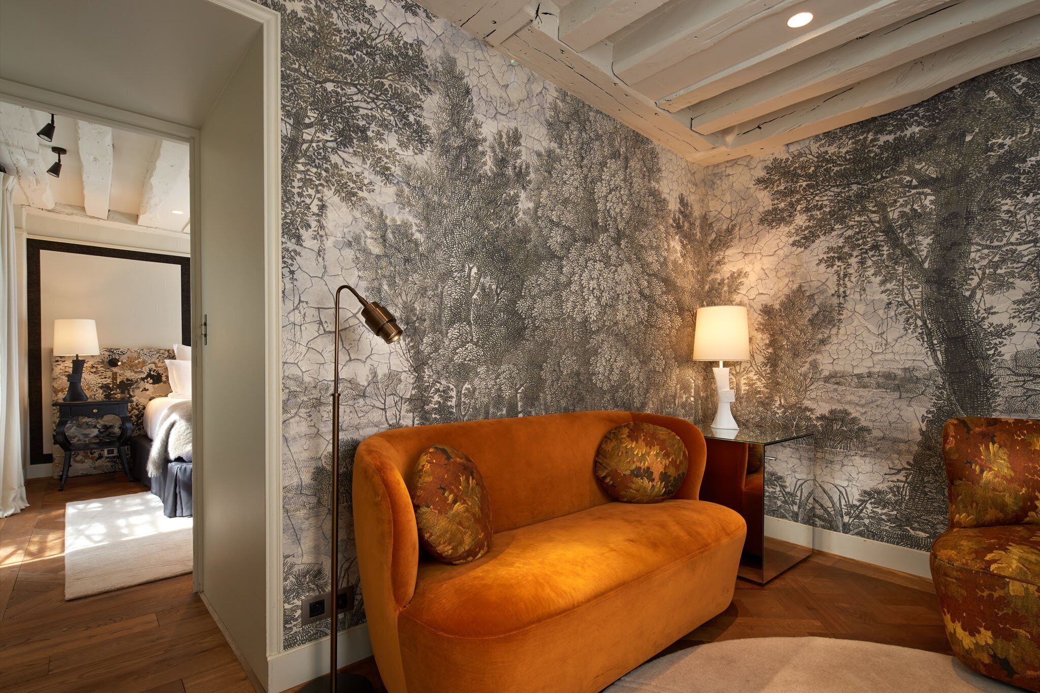 A room with a large mural of trees on the wall, a sofa with two decorative pillows, a floor lamp, and a small table with a lamp.