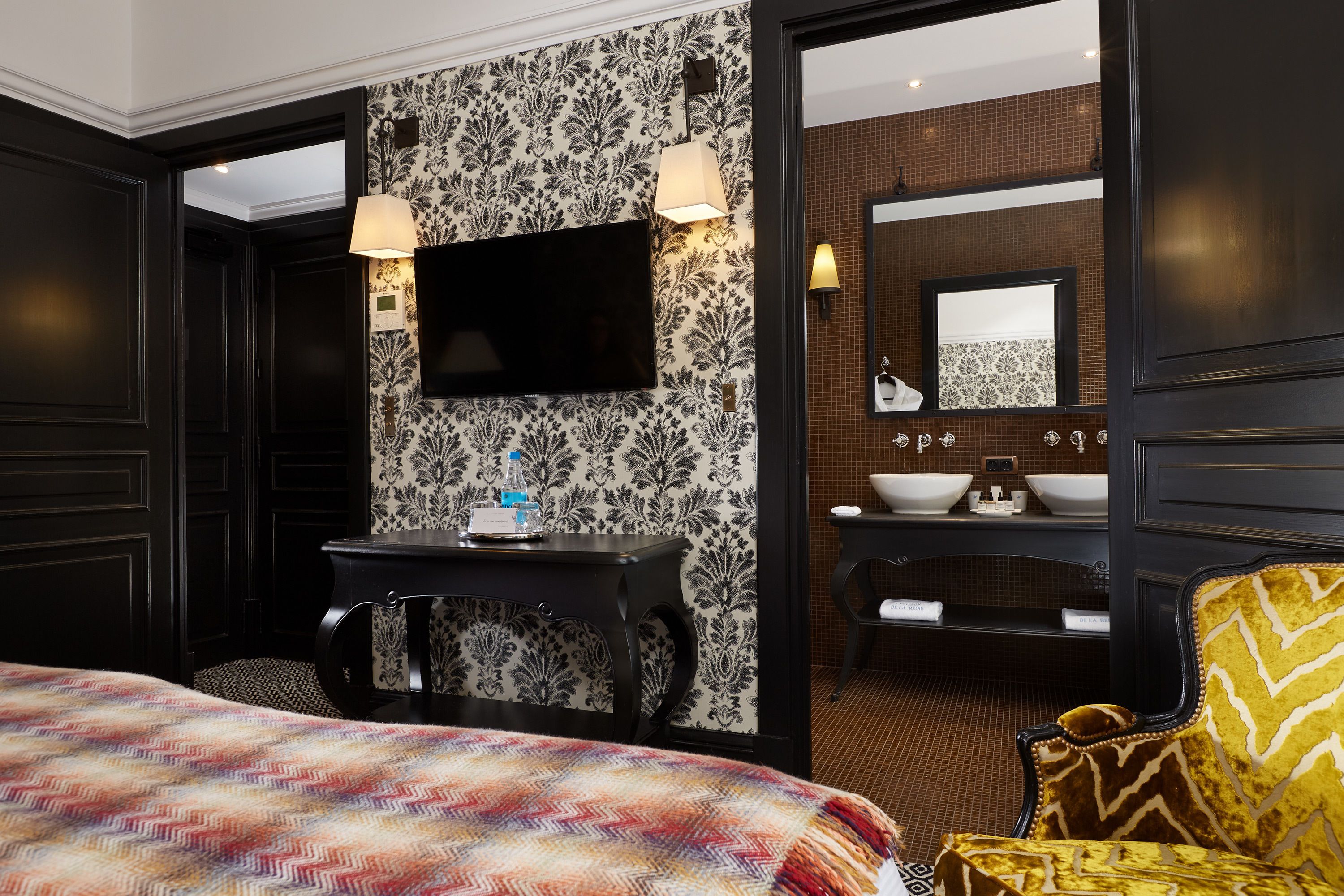 A hotel room with a bed, a desk with a flat-screen TV, a chair, and a bathroom with a sink and mirror.