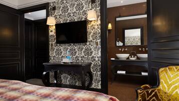 A hotel room with a bed, a desk with a flat-screen TV, a chair, and a bathroom with a sink and mirror.