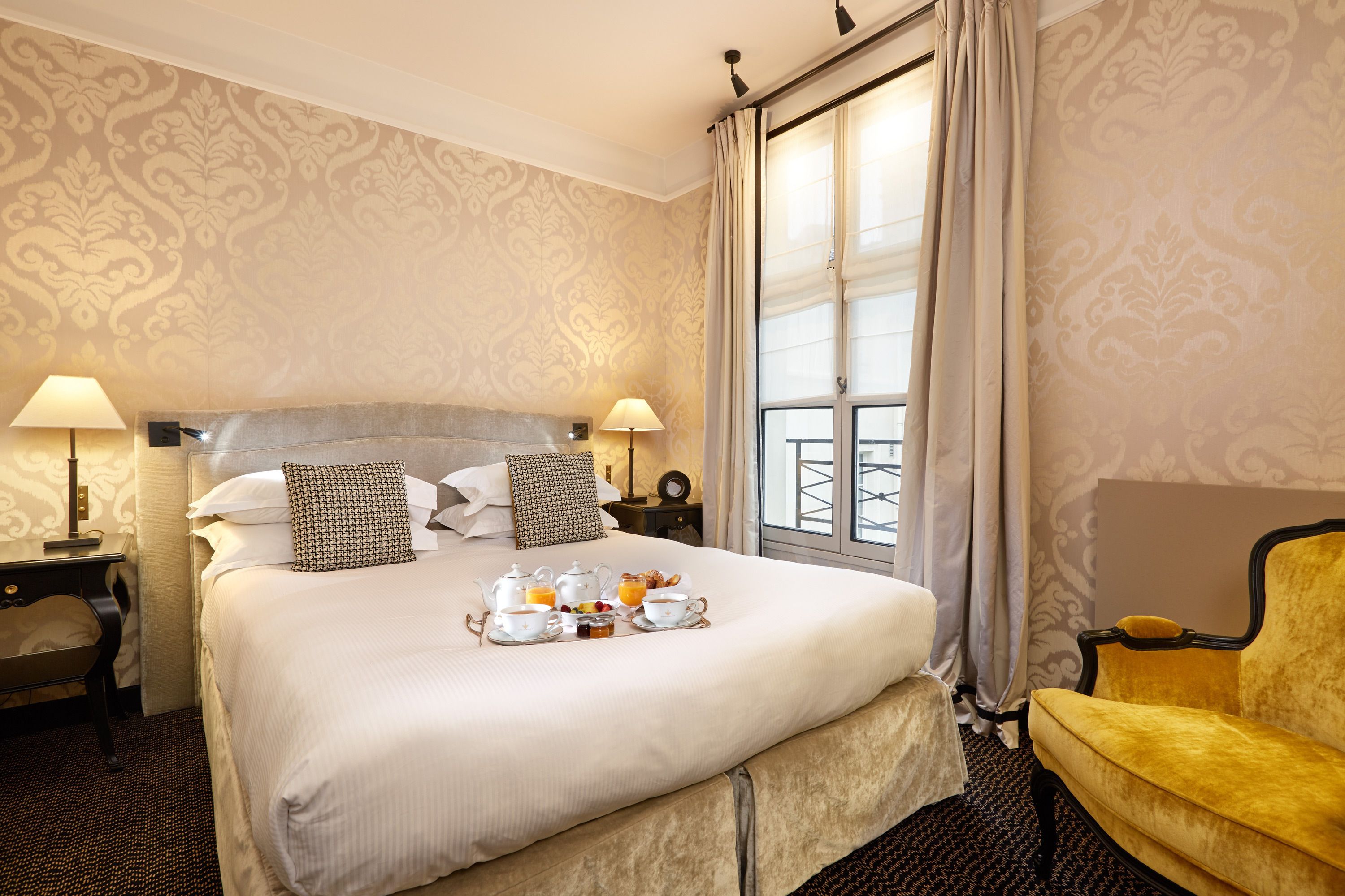 A hotel room with a large bed, two bedside lamps, a window with curtains, a yellow armchair, and a tray of breakfast items on the bed.