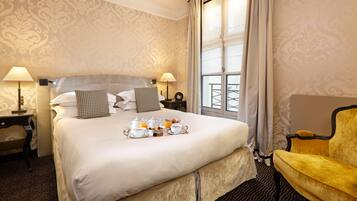 A hotel room with a large bed, two bedside lamps, a window with curtains, a yellow armchair, and a tray of breakfast items on the bed.