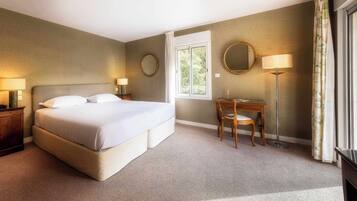 A hotel room with a large bed, bedside tables with lamps, a wooden chair, a desk, and a window with a view of greenery.