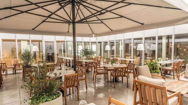 An indoor dining area with tables and chairs, large windows, and potted plants.