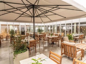 An indoor dining area with tables and chairs, large windows, and potted plants.