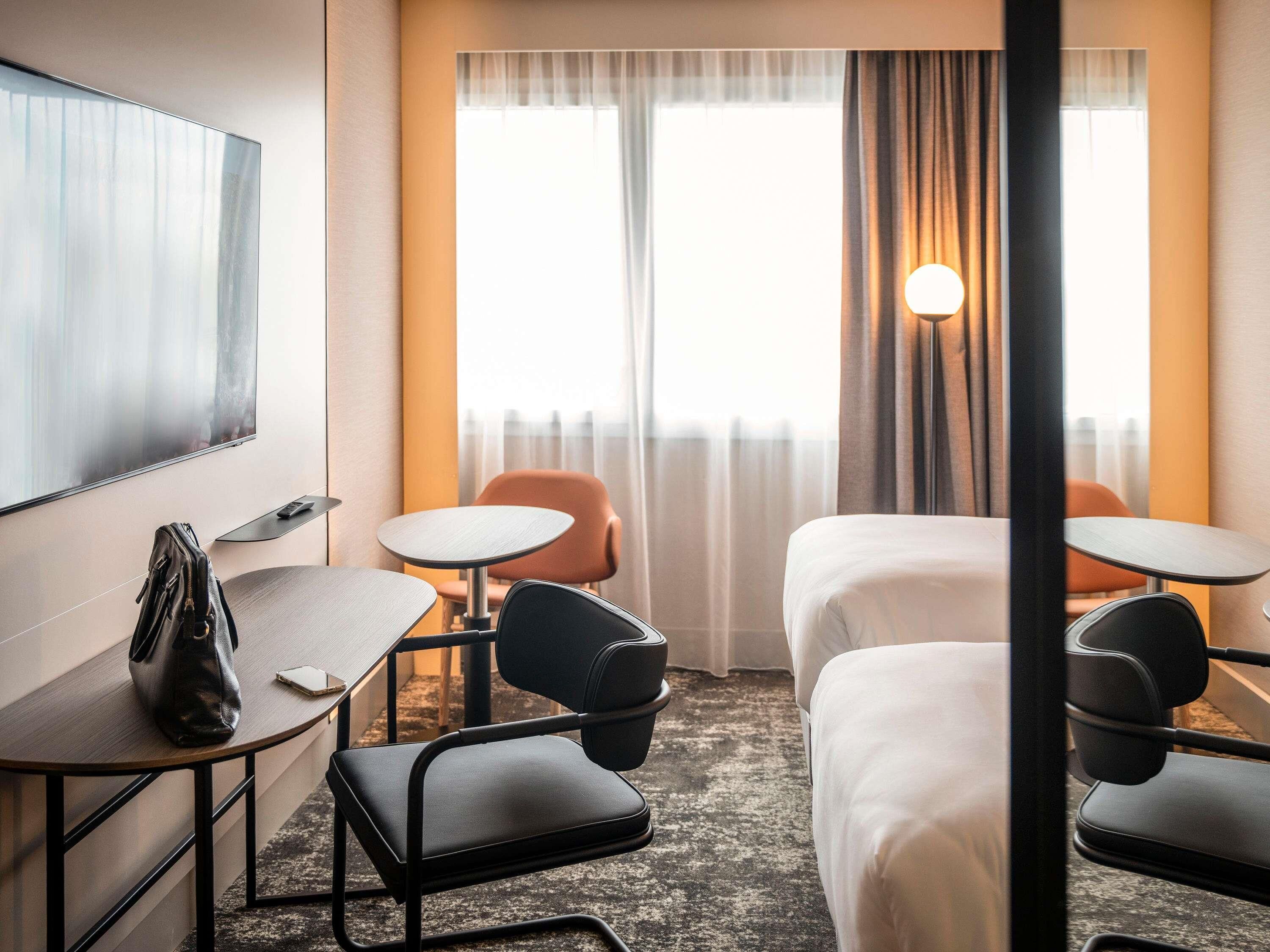 A hotel room with two beds, a desk, a chair, a TV, and a window with curtains.