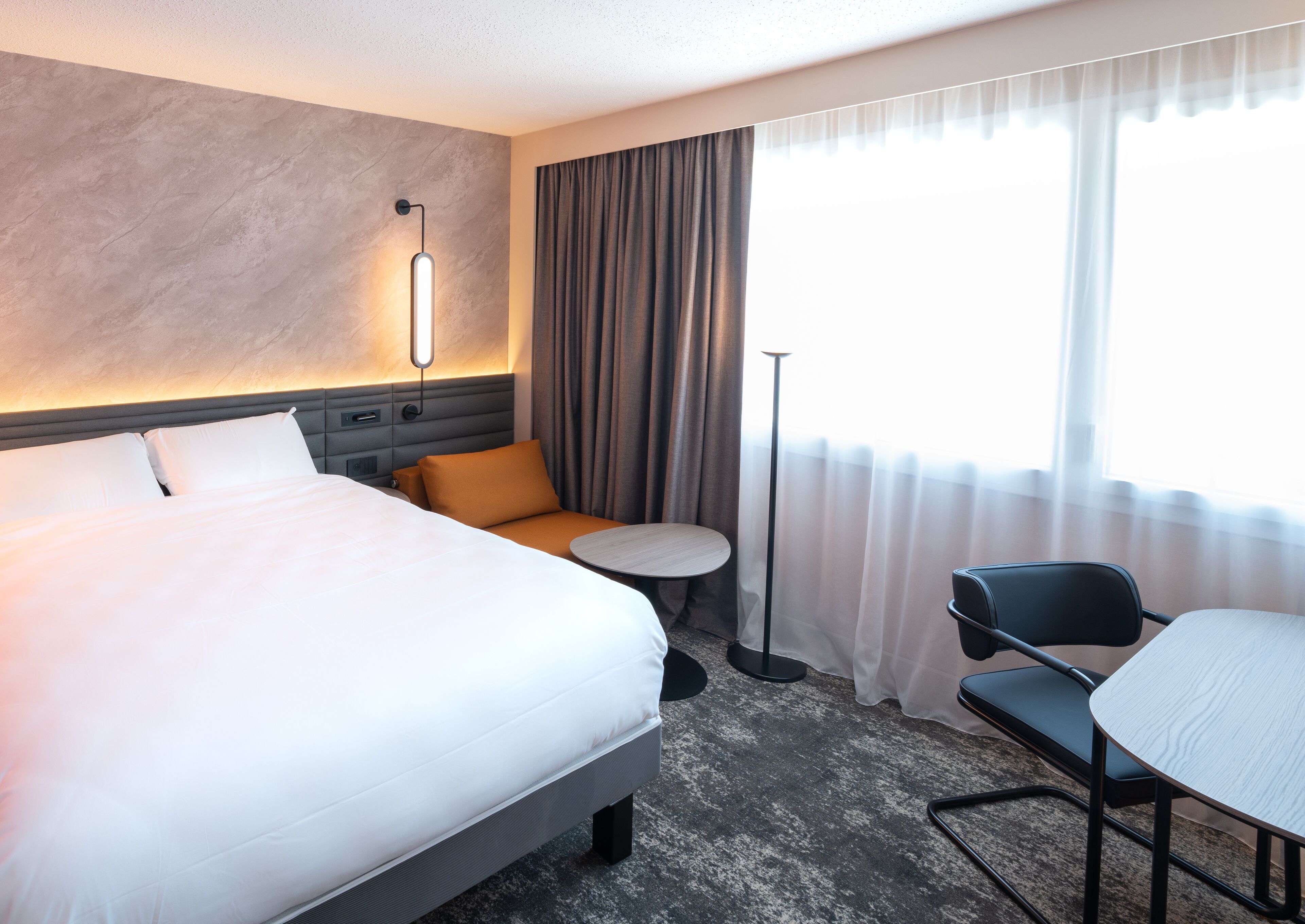 A hotel room with a large bed, a small round table, a chair, a desk, and a floor lamp.