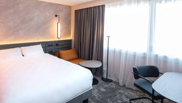 A hotel room with a large bed, a small round table, a chair, a desk, and a floor lamp.