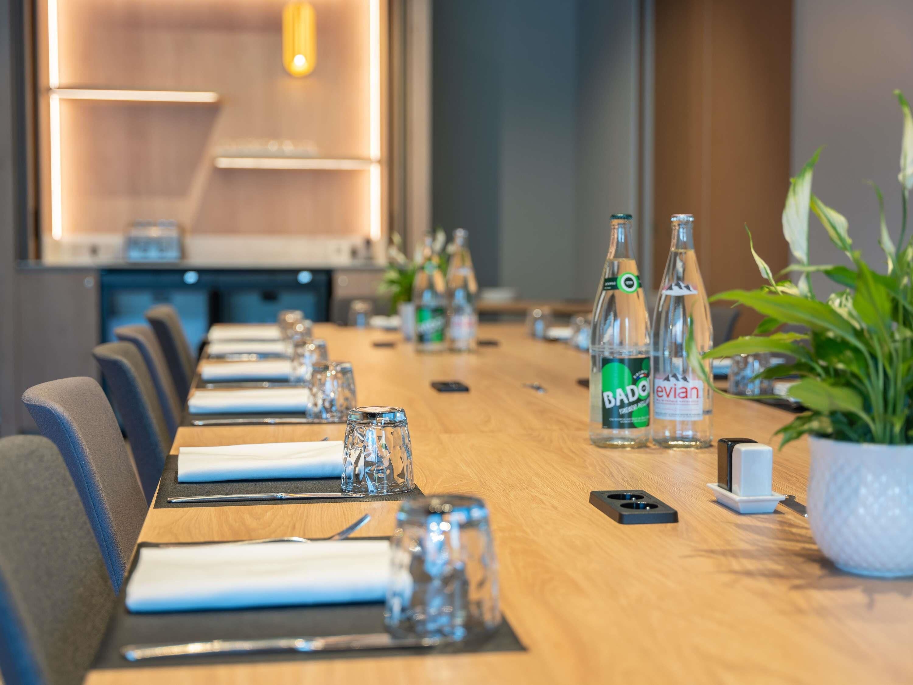 Een vergaderzaal met een lange tafel gedekt voor een vergadering, voorzien van glazen, waterflesjes en een potplant.