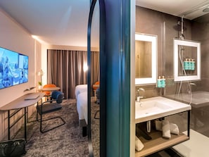 A hotel room with a bed, a desk with a chair, a TV, a shower area, and a bathroom with a sink and mirror.
