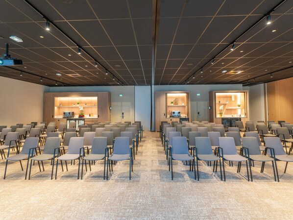A conference room with rows of chairs, a projector, and two reception areas with counters and plants.