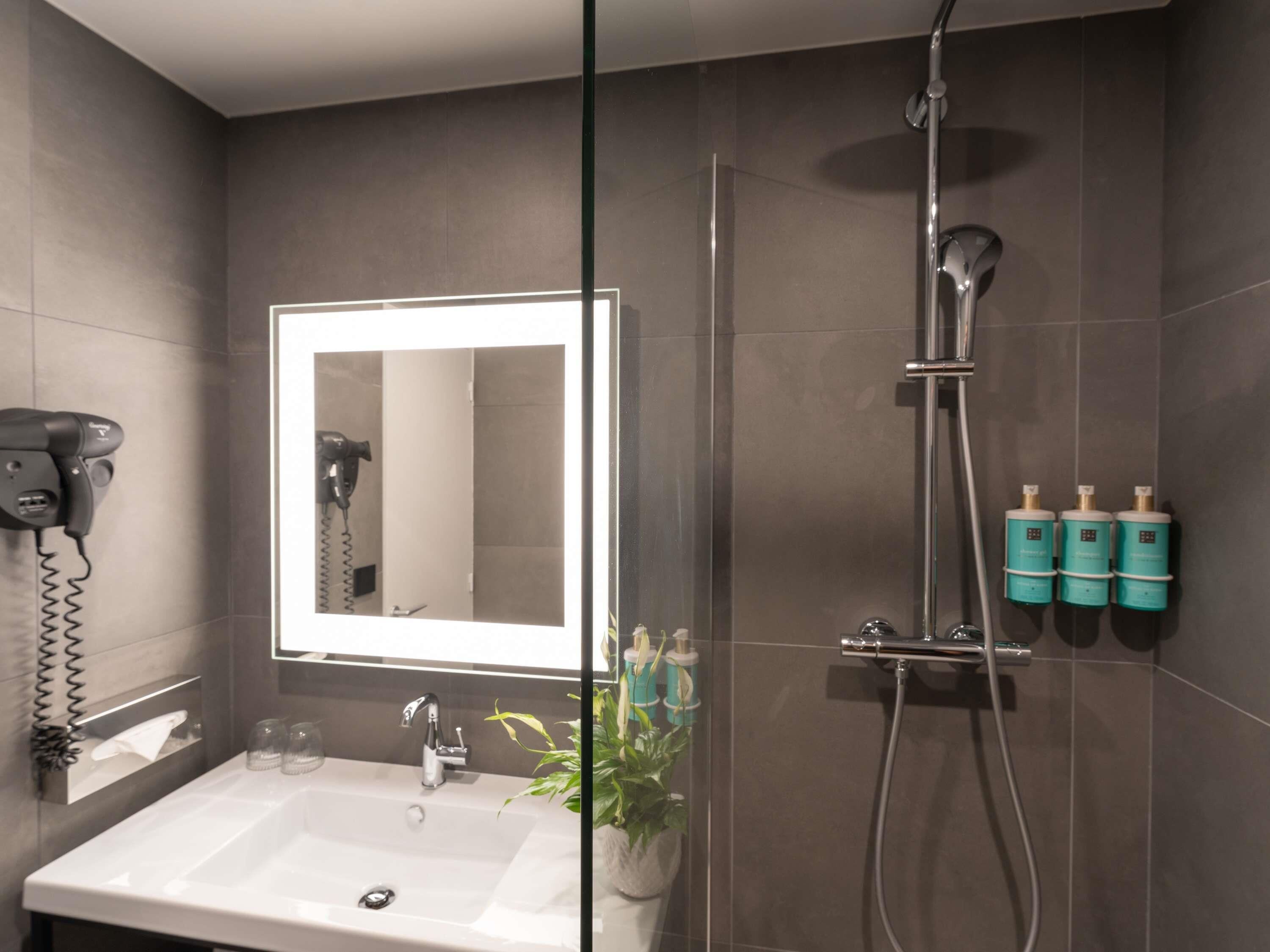 Ein Badezimmer mit Waschbecken, Spiegel, Fön, Dusche und Pflegeprodukten.