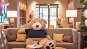 A plush bear sitting on a sofa in a well-lit lobby with a large window, a small round table, and a vase with dried flowers.