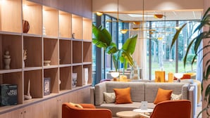 A modern hotel room with a wooden shelving unit, a grey sofa with orange cushions, and a round wooden table.