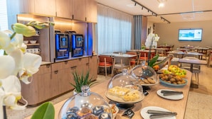A hotel dining area with a coffee machine, buffet with covered desserts, and a television screen.