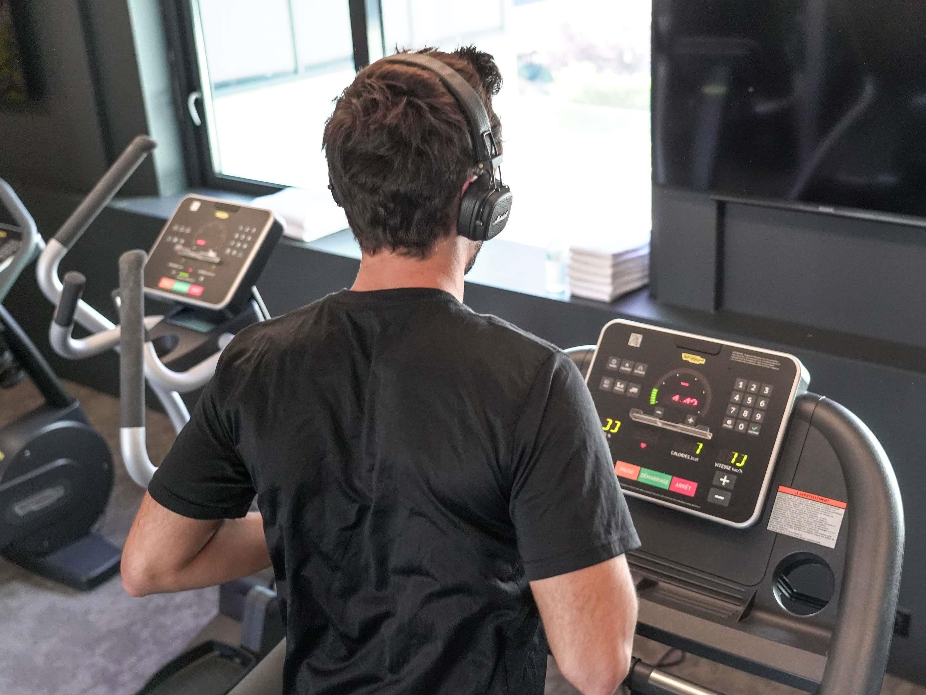 Une personne utilise un vélo d’appartement doté d’un écran numérique dans une salle de sport où l’on aperçoit, à l’arrière-plan, des tapis de course et des poids.