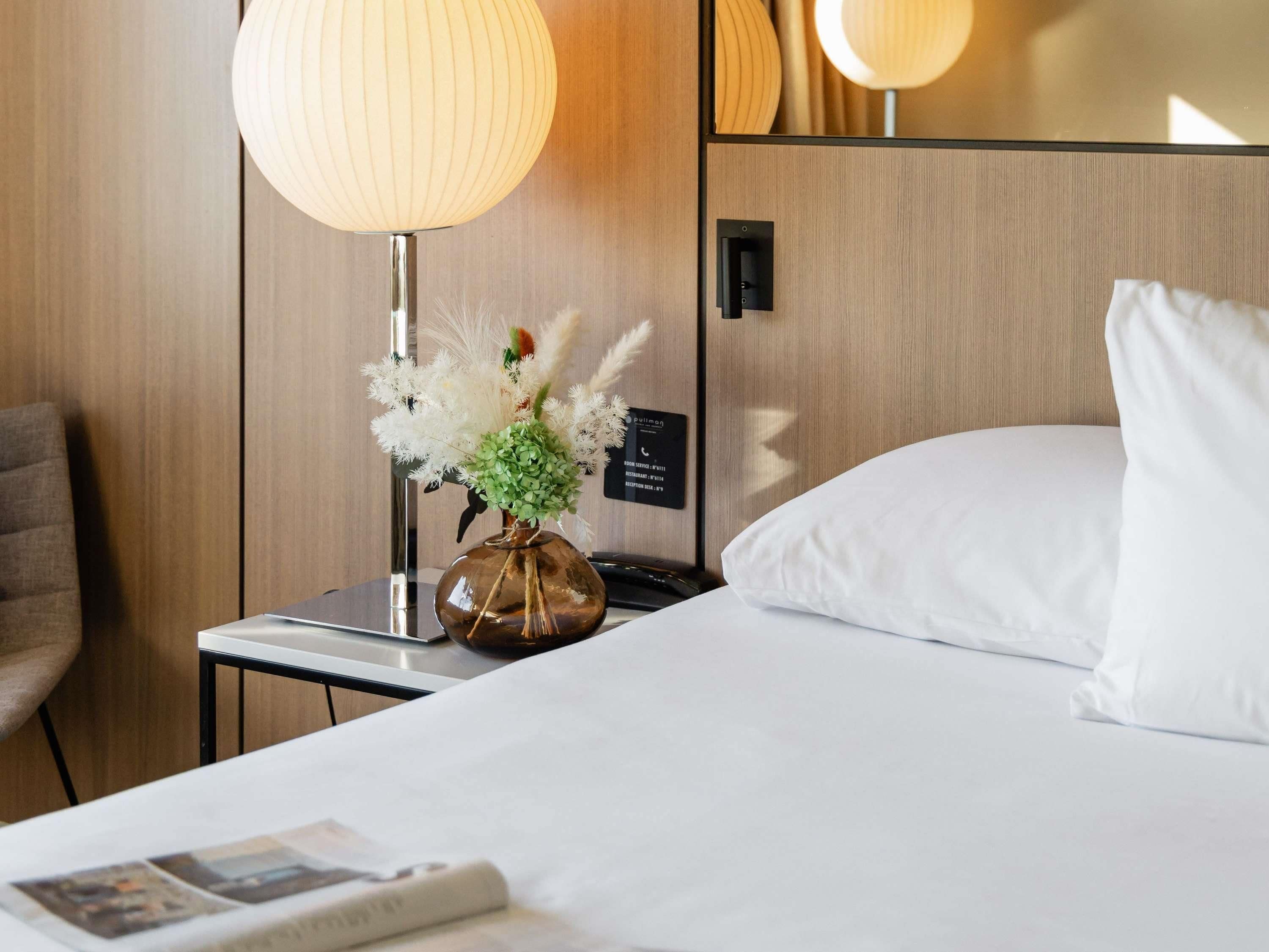 A hotel room with a bed, a nightstand with a lamp, a vase with flowers, and a wall-mounted phone.
