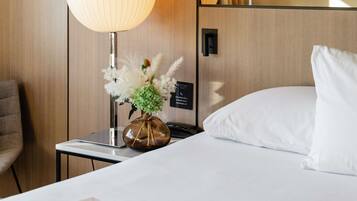 A hotel room with a bed, a nightstand with a lamp, a vase with flowers, and a wall-mounted phone.