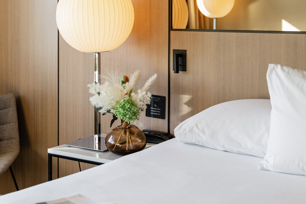 Una habitación de hotel con una cama, una mesita de noche con lámpara y un jarrón con flores.
