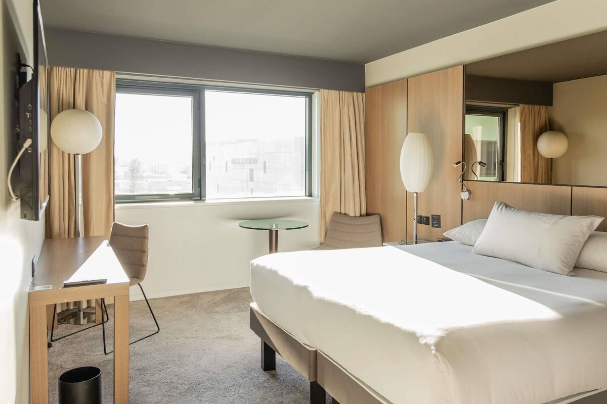 A hotel room with a bed, a desk, a chair, a small table, a lamp, and a window with curtains.