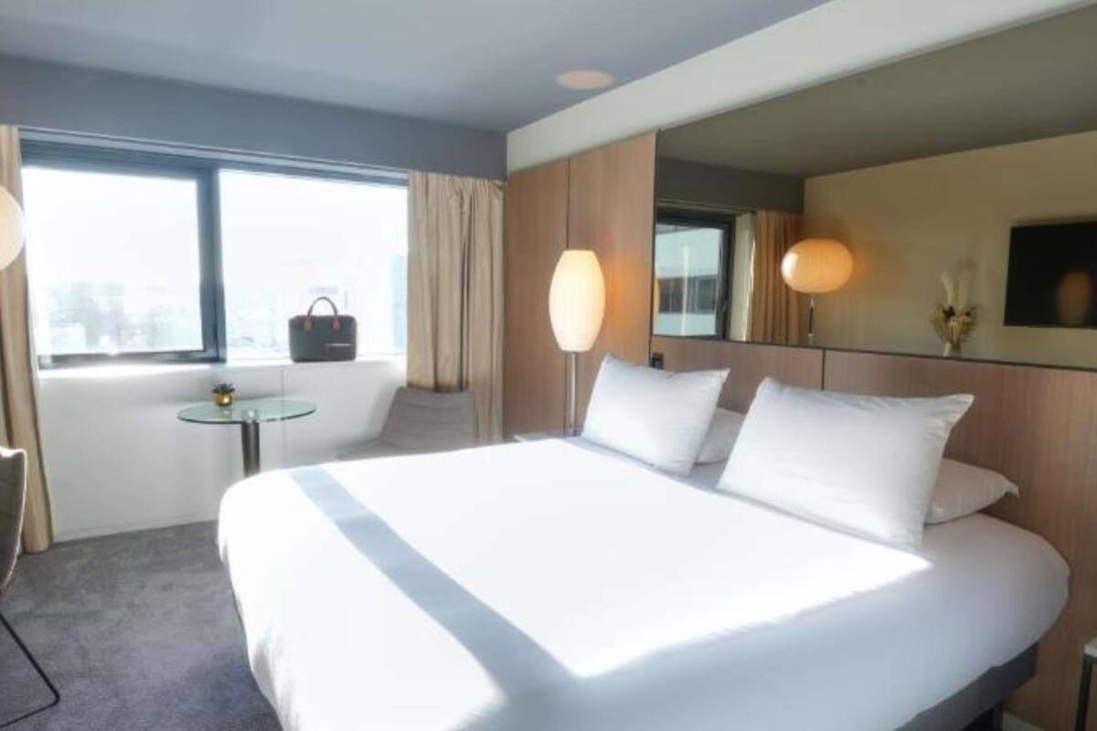 A hotel room with a large bed, a small table, a chair, a window, and a mirror.