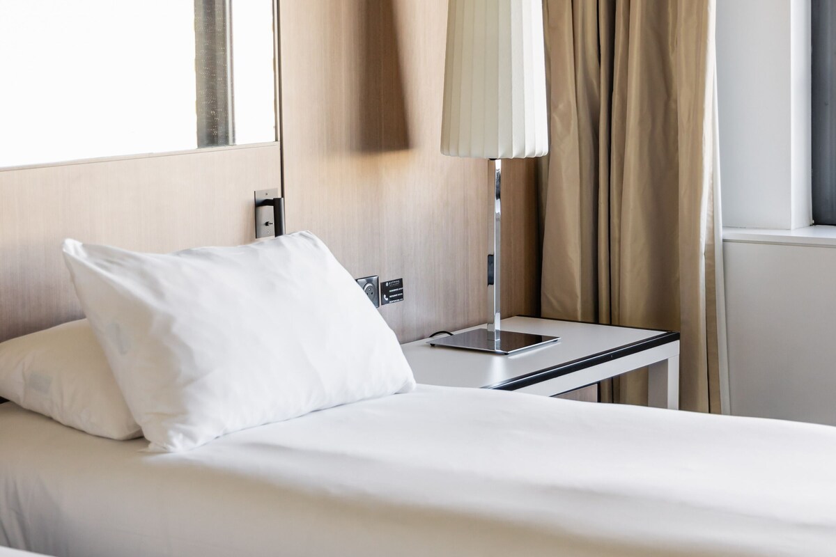 Una cama bien tendida con sábanas blancas, una mesita de noche con una lámpara y una ventana con cortinas.