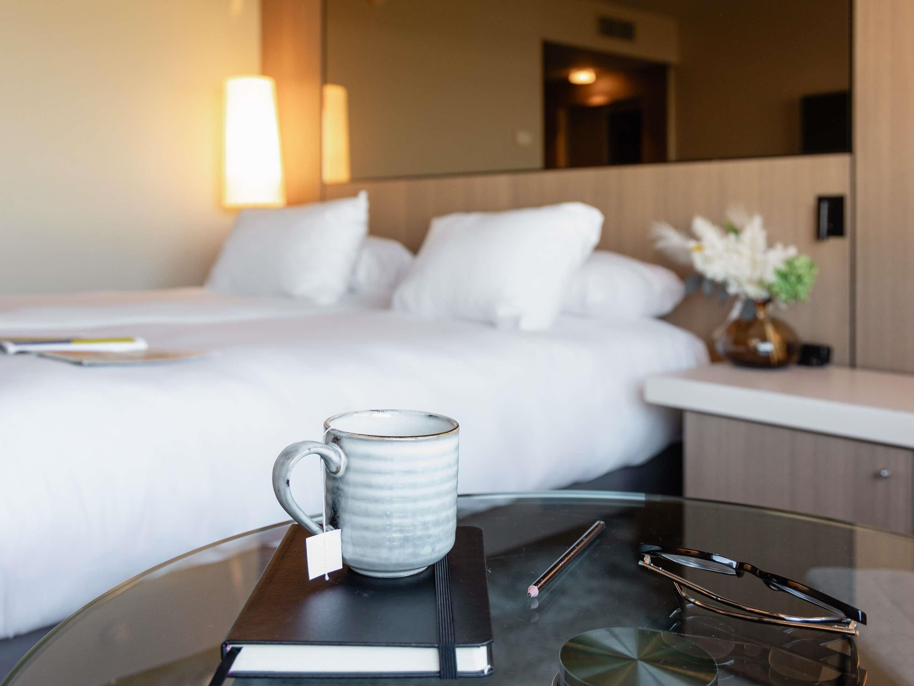 A hotel room with a bed, a bedside table with a mug, glasses, and a book.