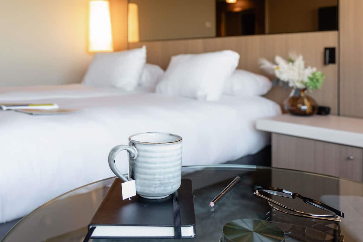 A hotel room with a bed, a bedside table with a mug, glasses, and a book.