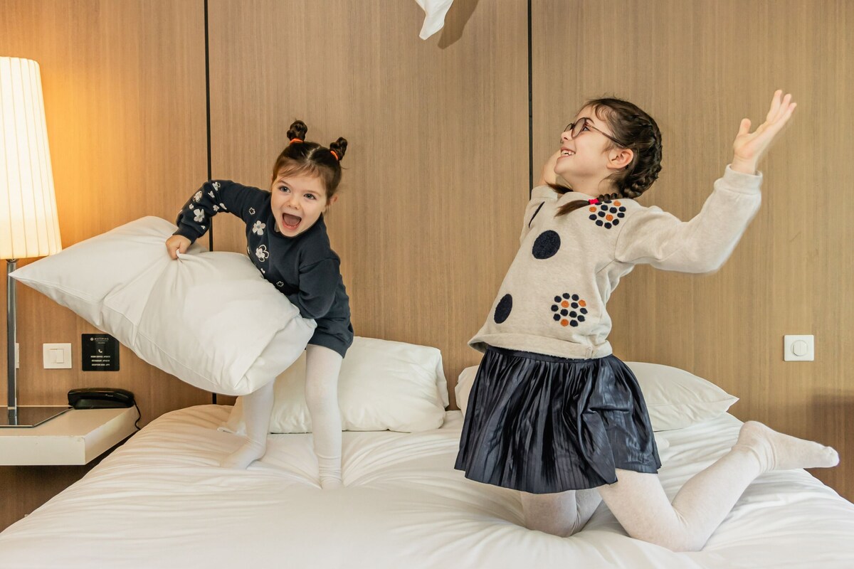 Dos niños jugando en una cama dentro de una habitación de hotel con paneles de madera y una mesita de noche con lámpara.