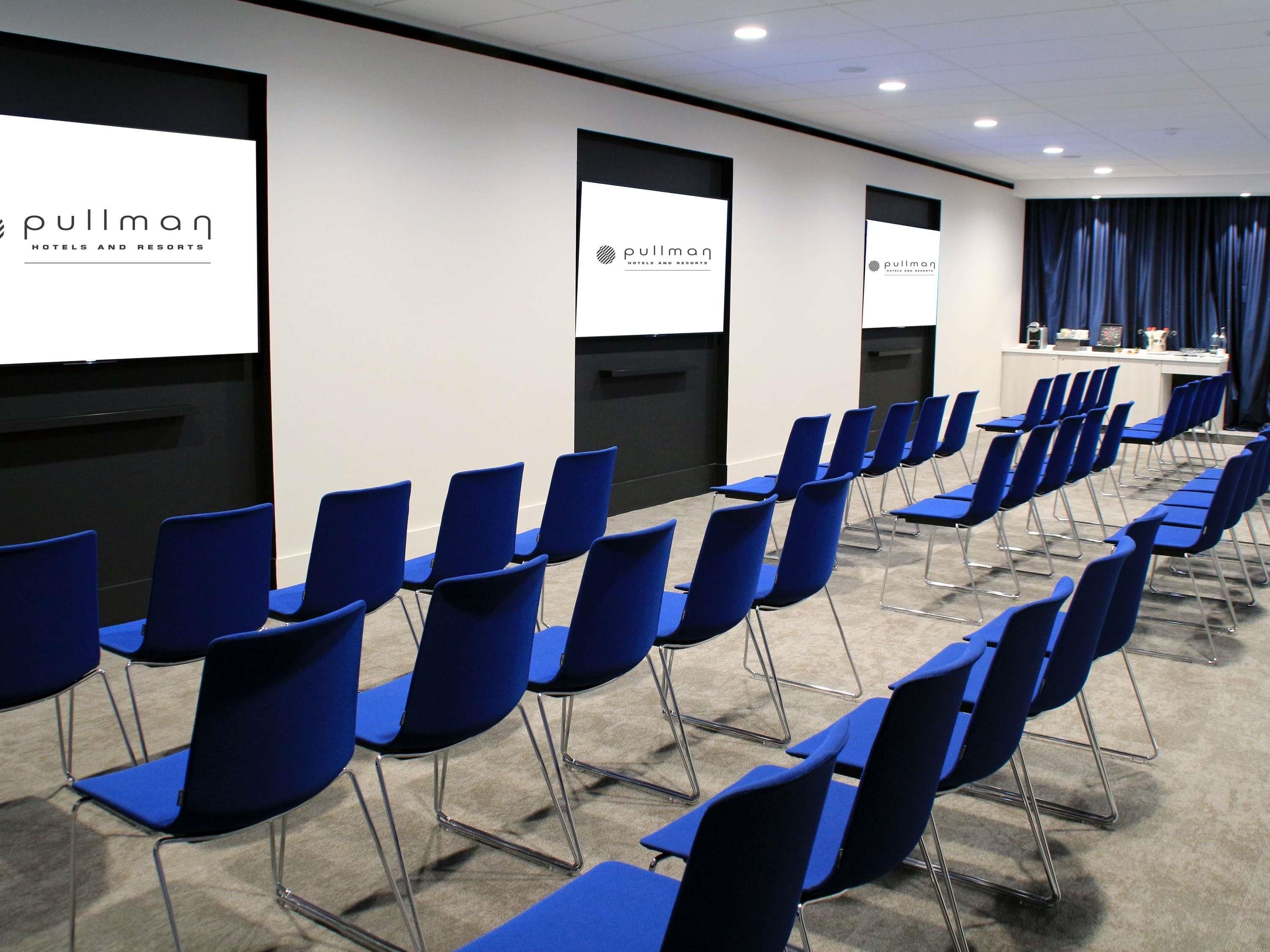 Un salón de conferencias con filas de sillas azules, un atril y una pantalla que muestra el logotipo de Pullman Hotels and Resorts.