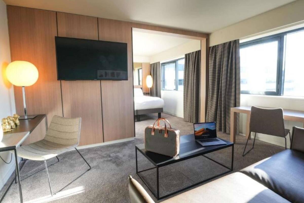 A hotel room with a bed, a desk with a laptop, a TV, and a view of the city.