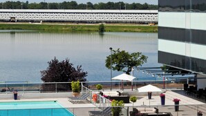 A swimming pool with lounge chairs, a tree, and a view of a large building across the water. - Hotel Pullman Bordeaux Lac (Bordeaux)
