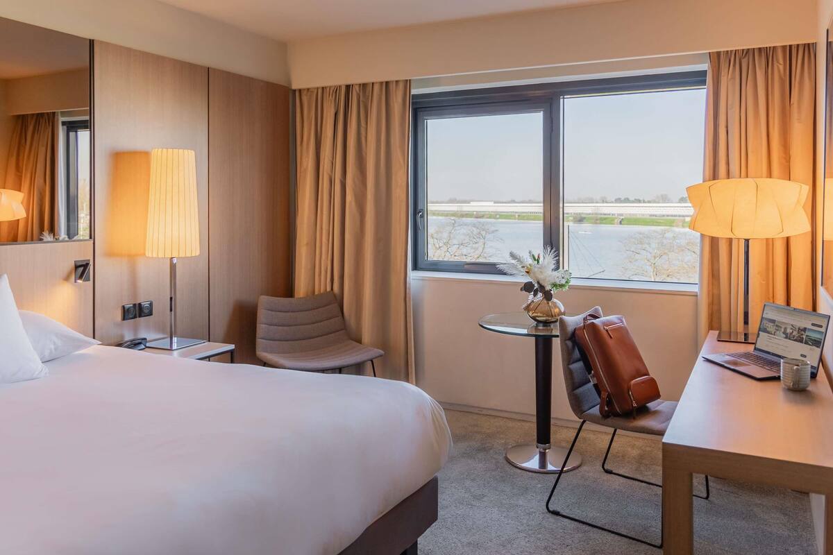 A hotel room with a bed, a desk, a chair, a lamp, a small table with a vase, and a view of a body of water through the window.