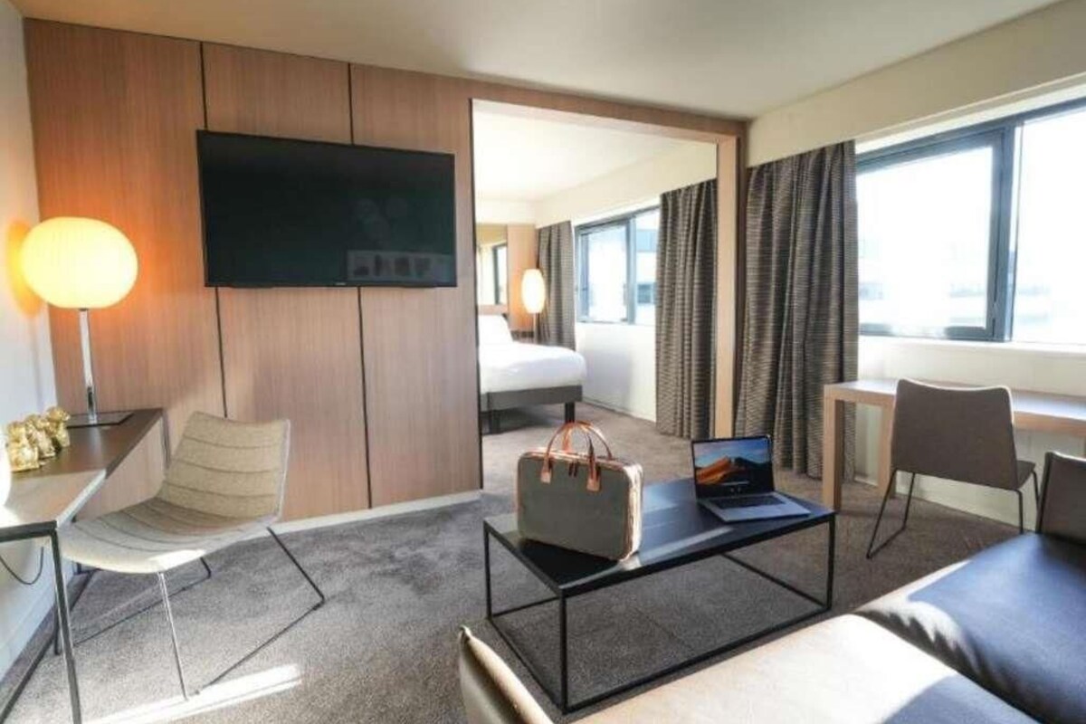 Habitación de hotel con televisión de pantalla plana, escritorio con silla, sofá, mesa de centro con bolso y cama con cortinas.