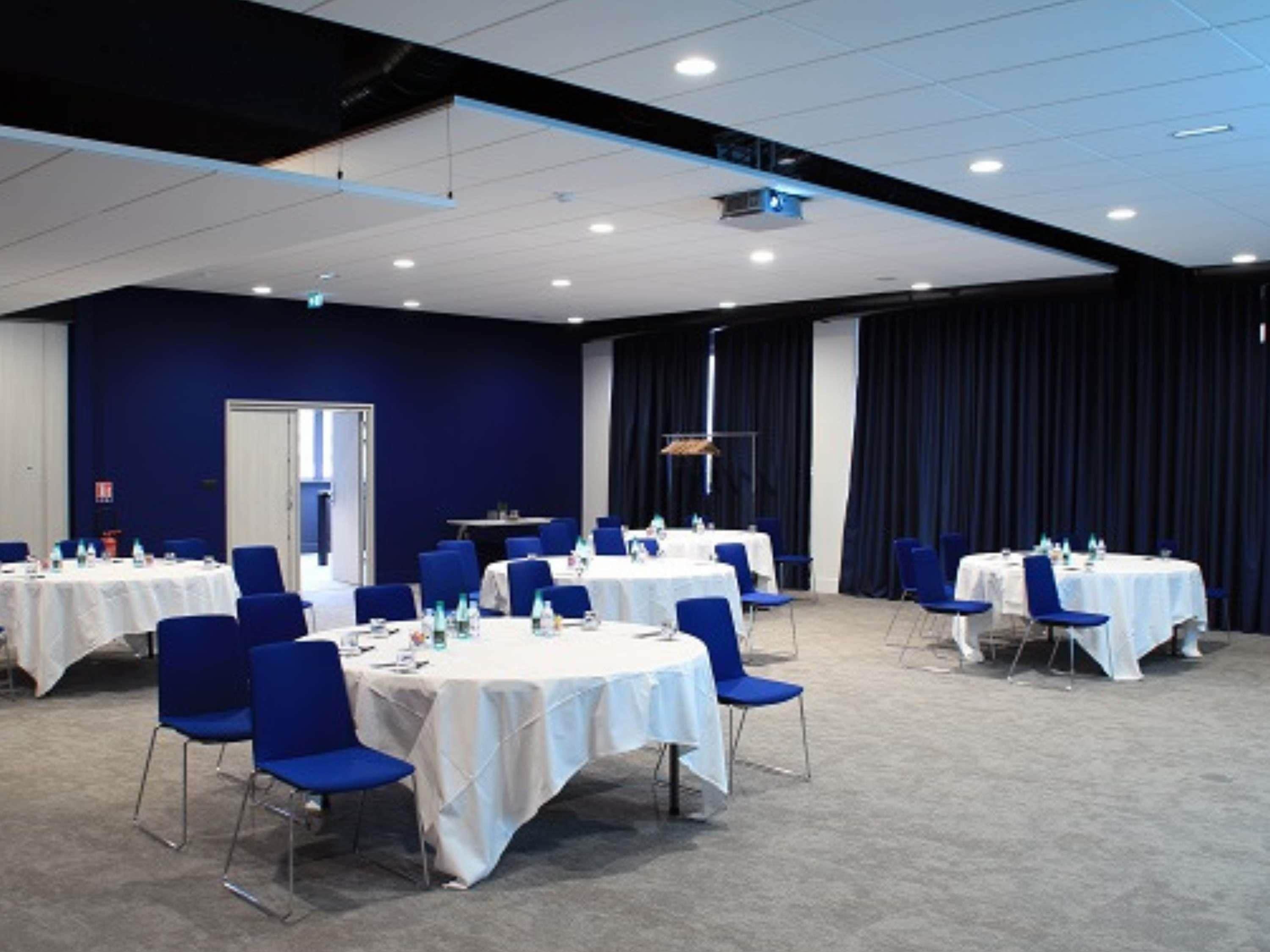 Un salón de conferencias con varias mesas y sillas, un escenario y un telón oscuro de fondo.