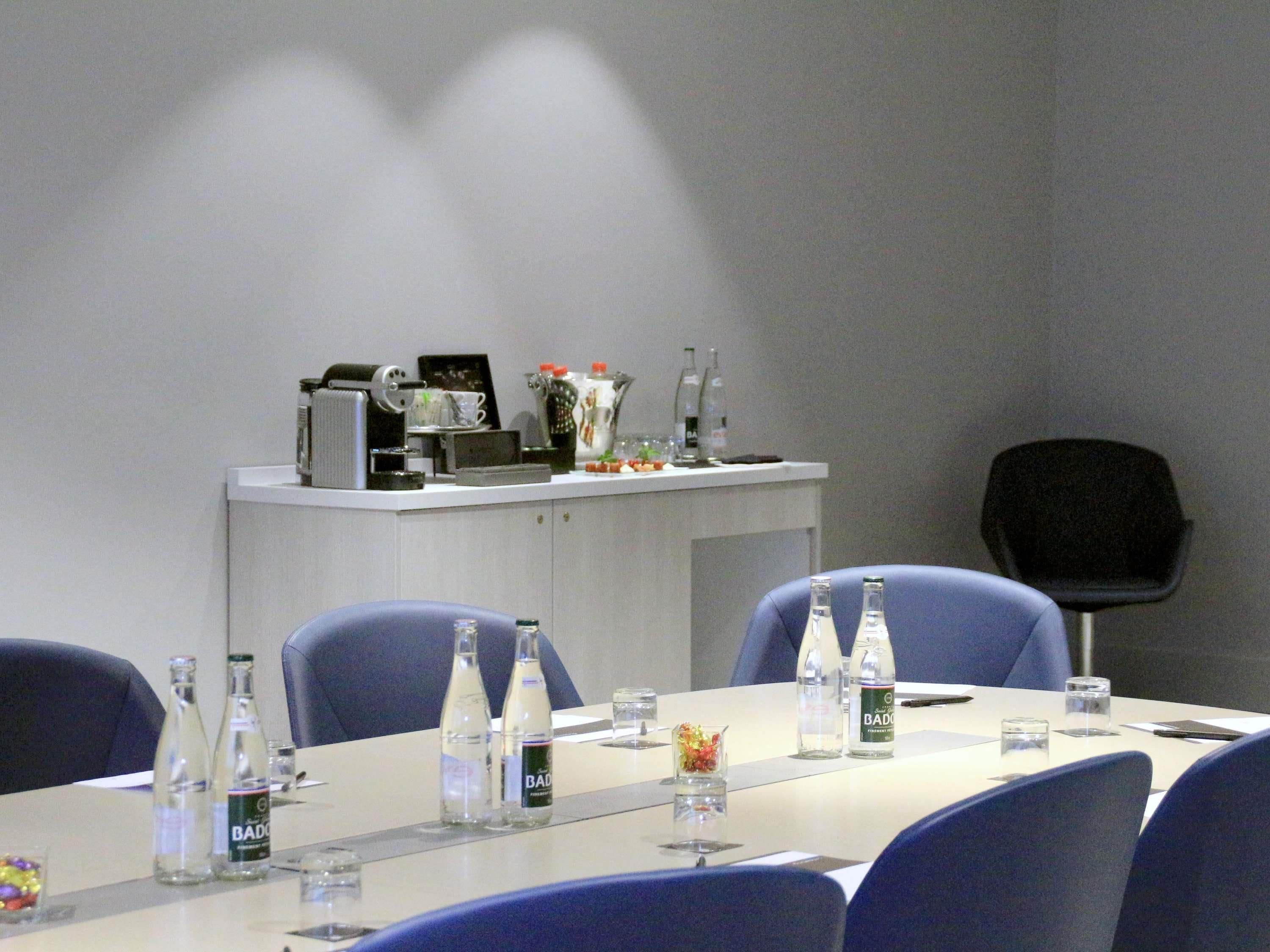 Ein Konferenzraum mit langem Tisch, Stühlen, Wasserflaschen und einer Kaffeestation.