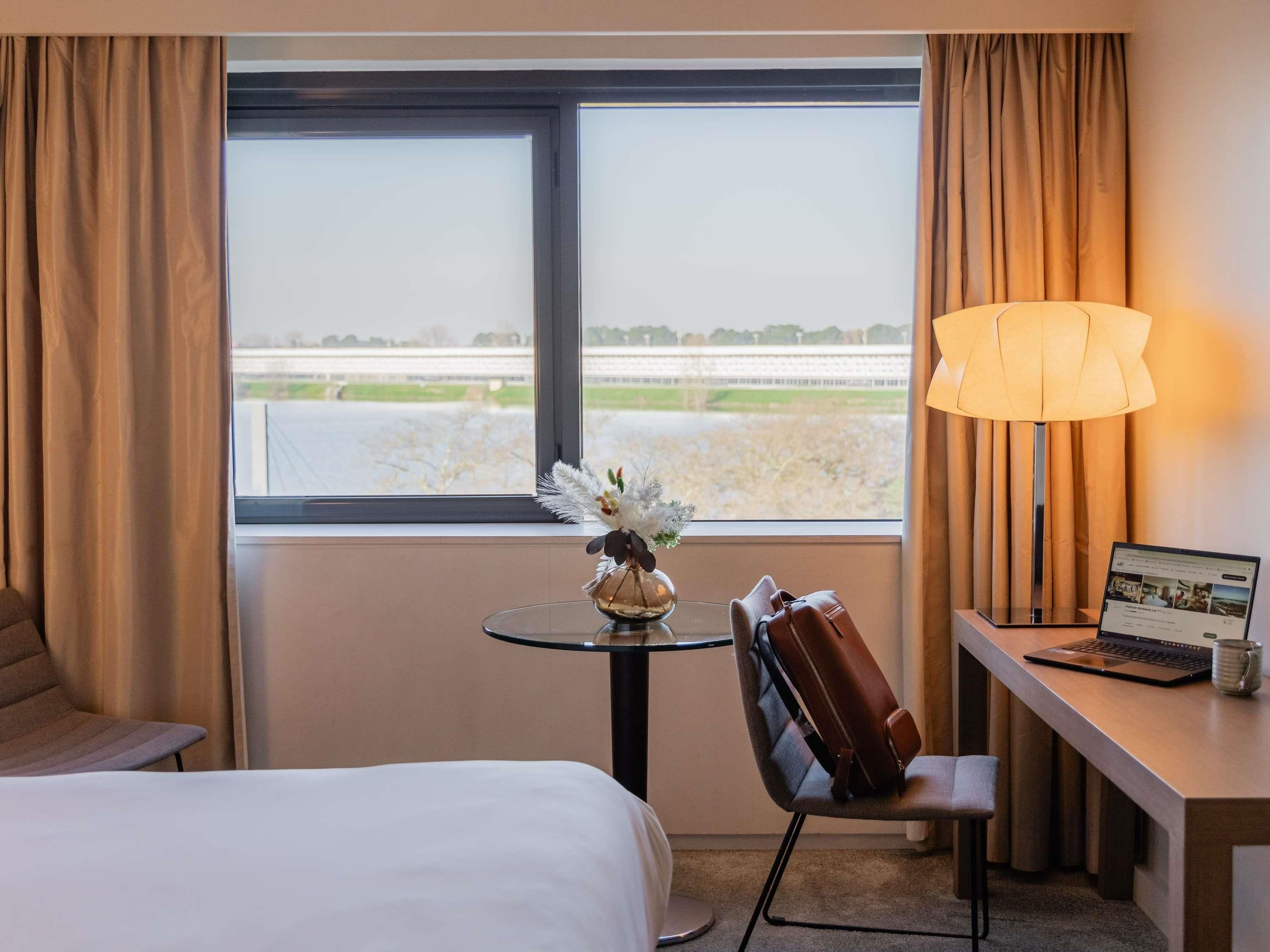 A hotel room with a bed, a desk with a laptop, a chair, a lamp, and a view of a bridge and greenery.