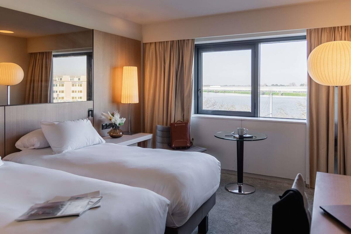 A hotel room with two beds, a small table, a chair, a lamp, and a view of a body of water.