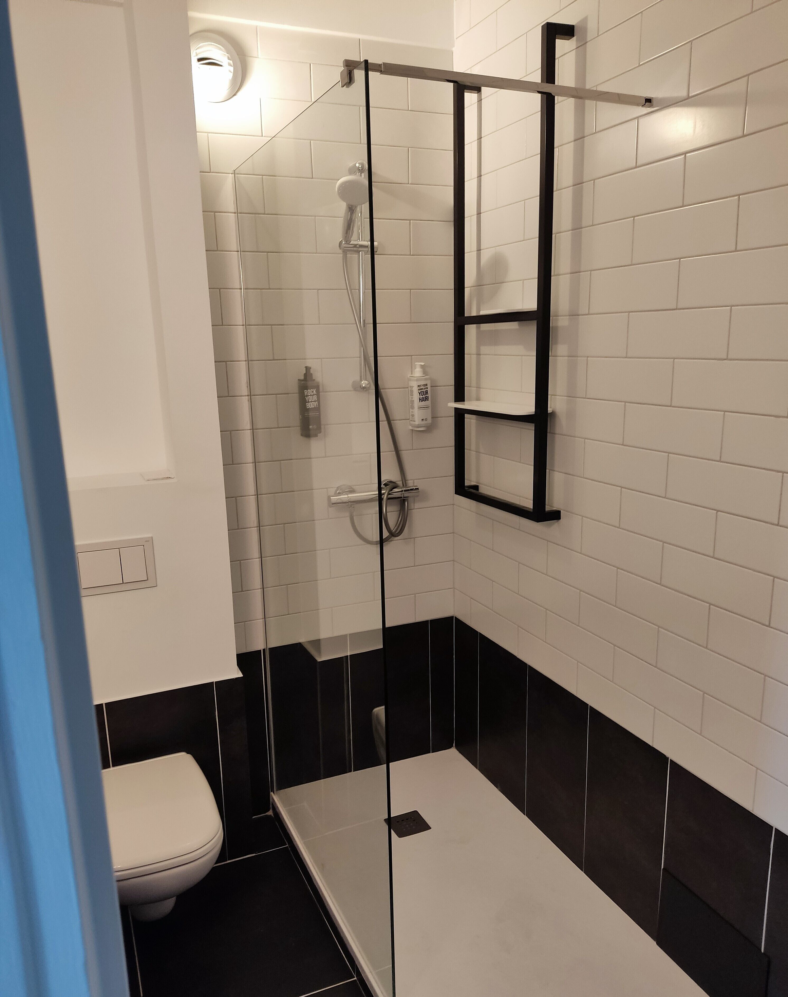 Un baño moderno con ducha de vidrio, paredes de azulejos blancos y negros, un inodoro y un estante con artículos de aseo.
