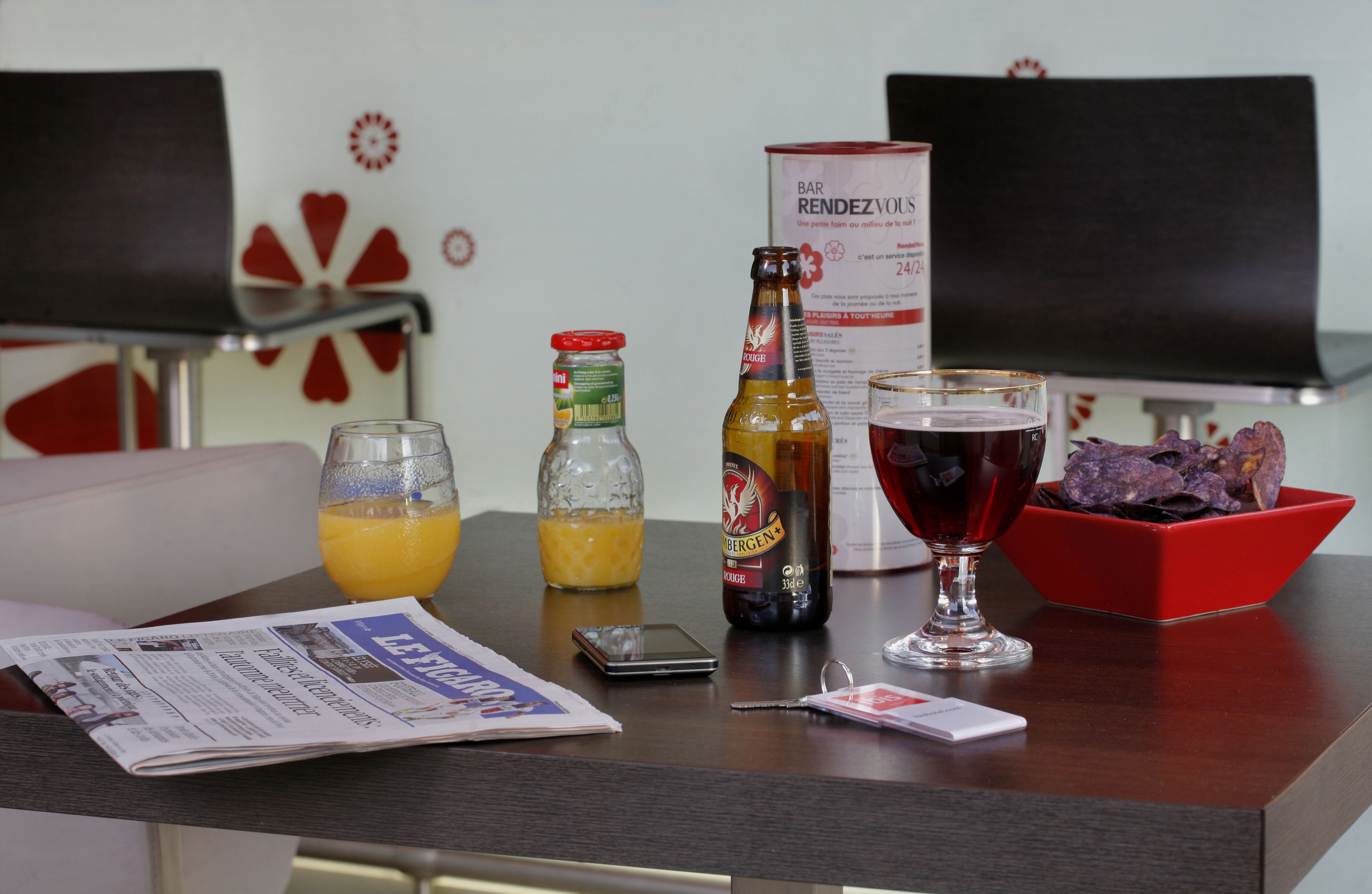 Une chambre d’hôtel avec une table à manger dressée, sur laquelle on trouve un journal, un verre de jus d’orange, une bouteille de bière, un verre de vin rouge, un bol de collations et un présentoir à menus.