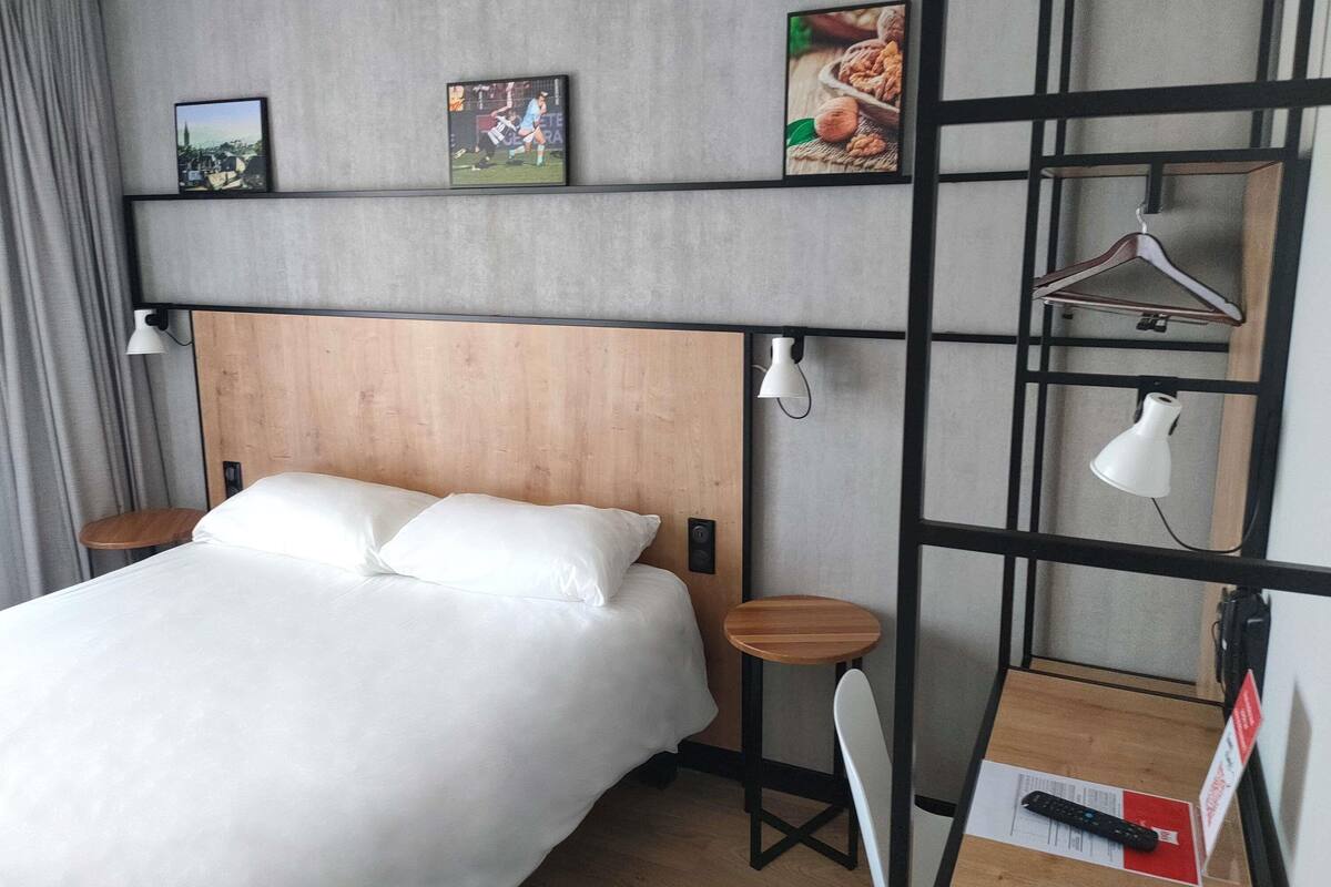 A hotel room with a bed, a desk, a chair, a hanging light, and a shelf with pictures.