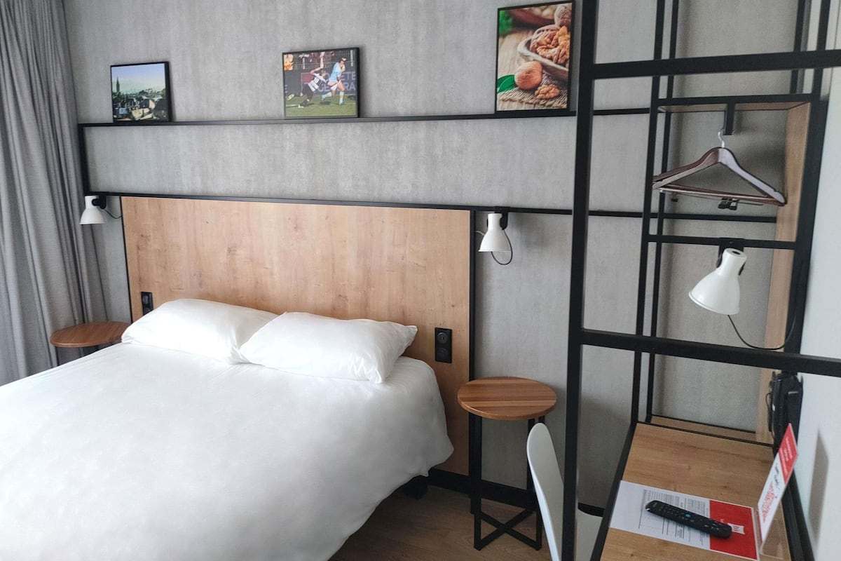 A hotel room with a bed, a desk, a chair, a hanging light, and a shelf with pictures.