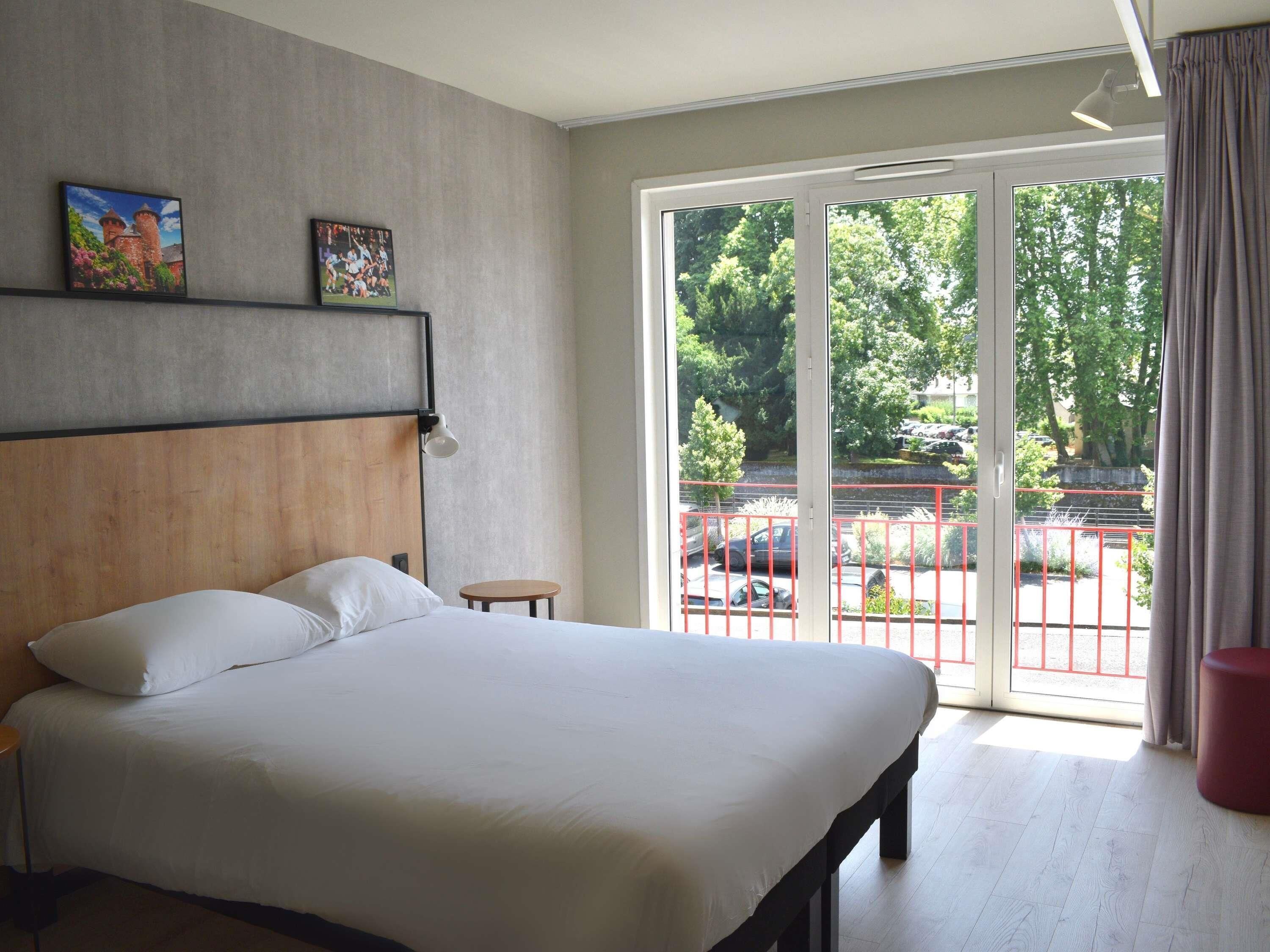 A hotel room with a bed, a small table, a nightstand, a wall-mounted lamp, and a large sliding glass door leading to an outdoor area with a view of trees and parked cars.