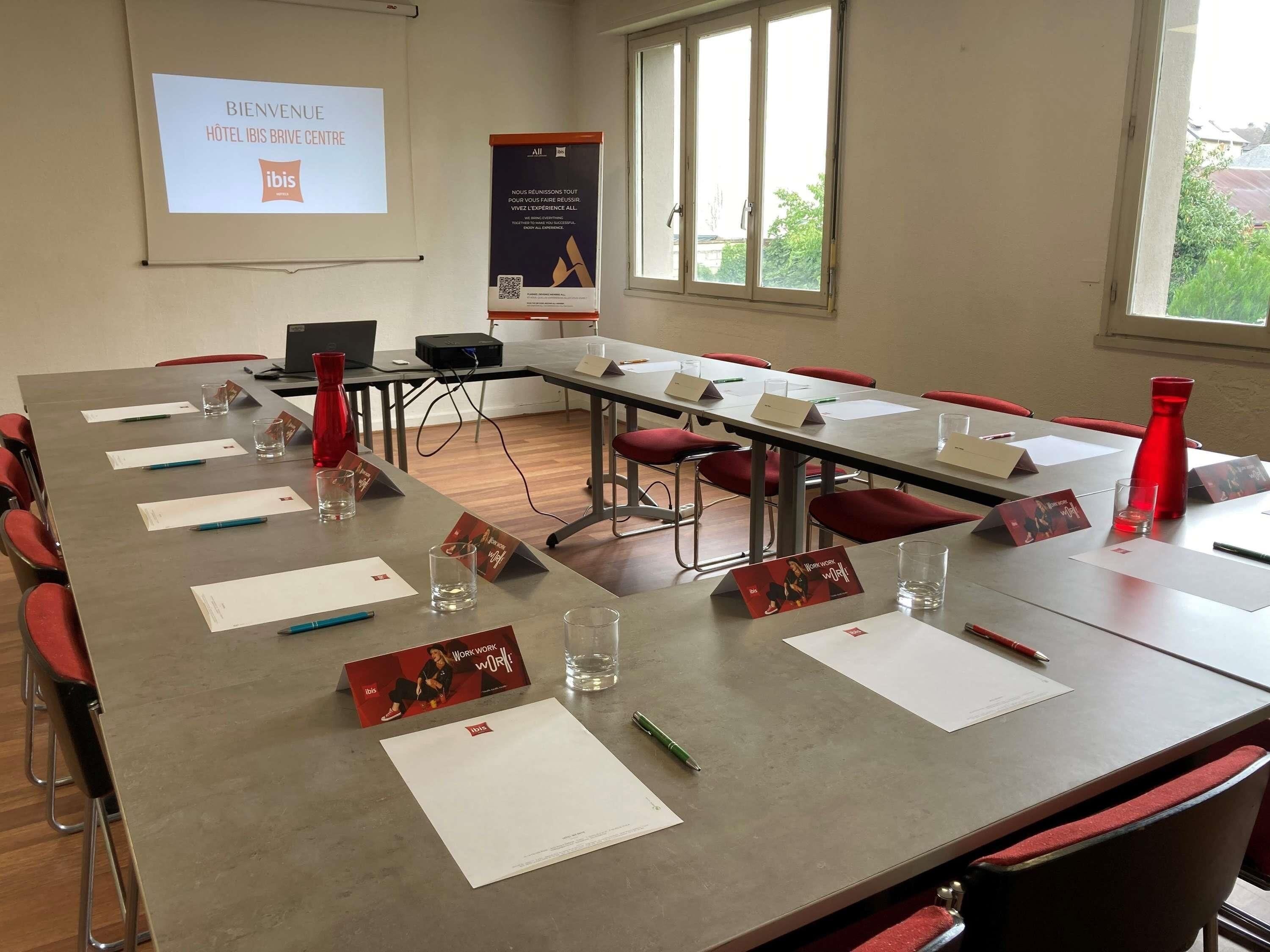 Uma sala de conferências com uma mesa comprida, cadeiras vermelhas, cartões de nome e uma tela de projetor exibindo 'BIENVENUE HOTEL IBIS BRIVE CENTRE'.