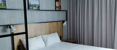 Una habitación de hotel con una cama, cabecera de madera, una mesita con una silla, una lámpara de noche y un cuadro en la pared.