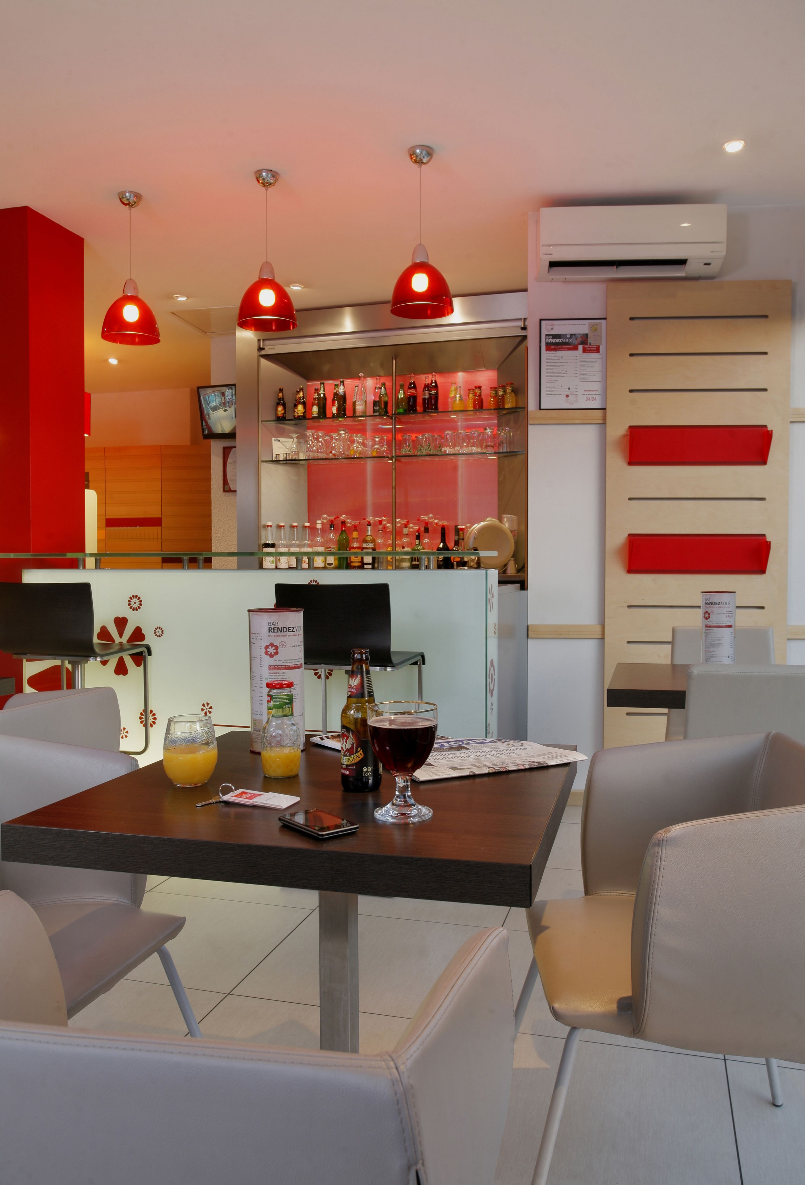 A modern bar area with a bar counter, red pendant lights, and a selection of bottles and glasses.