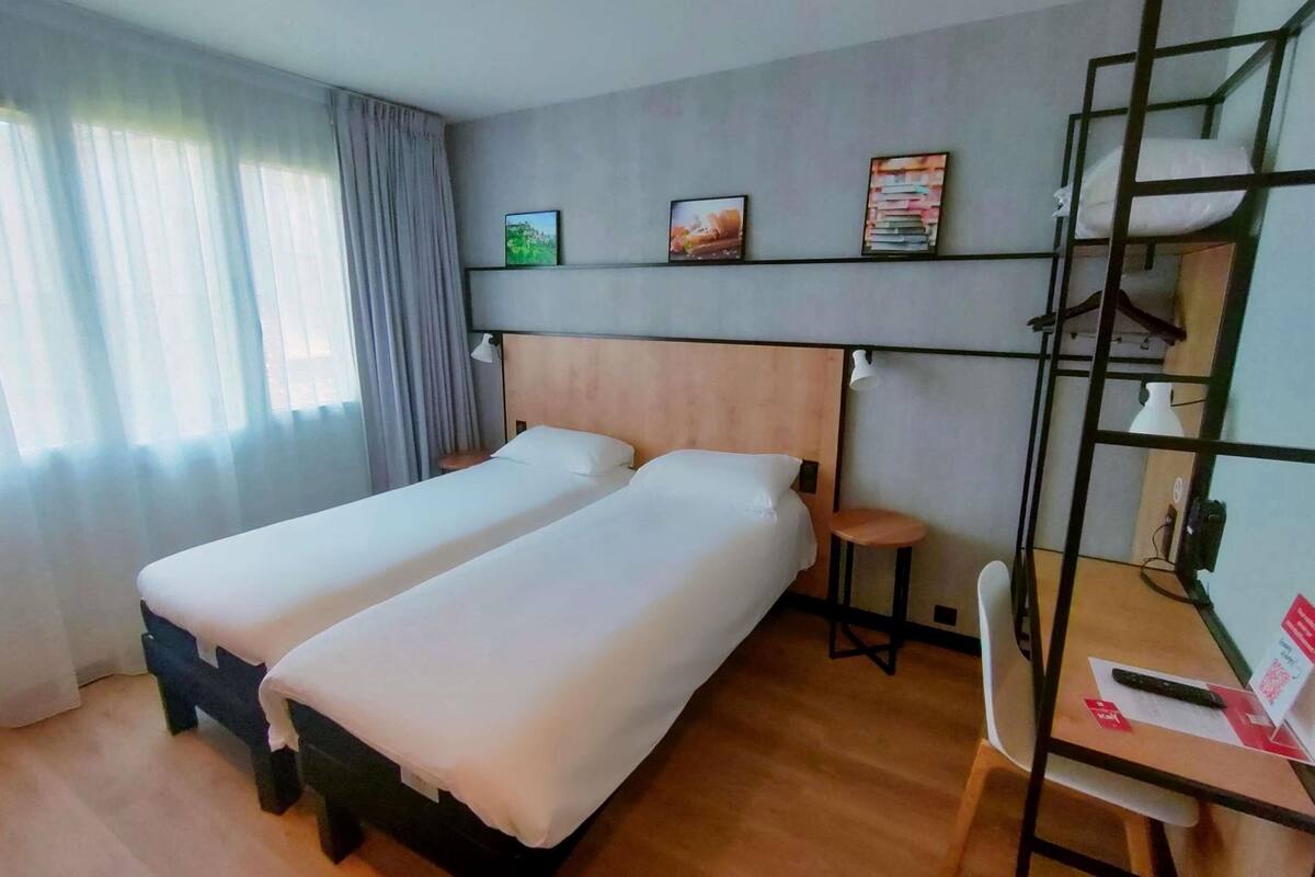 A hotel room with two beds, a desk, a chair, a shelf with books, and a window with curtains.