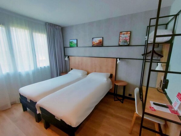 A hotel room with two beds, a desk, a chair, a shelf with books, and a window with curtains.