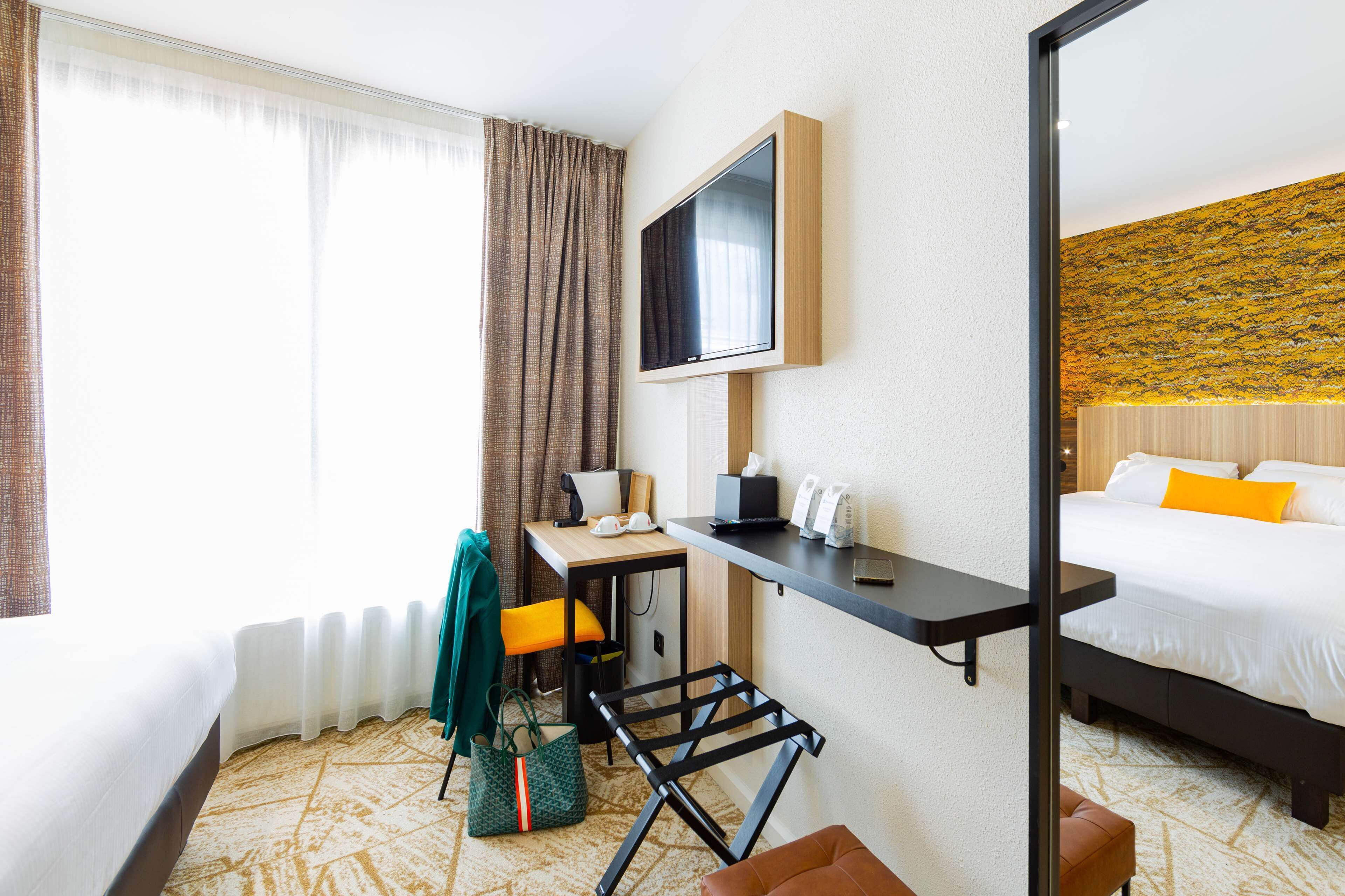 A hotel room with a bed, a desk with a computer, a chair, a small table, a TV mounted on the wall, and a window with curtains.