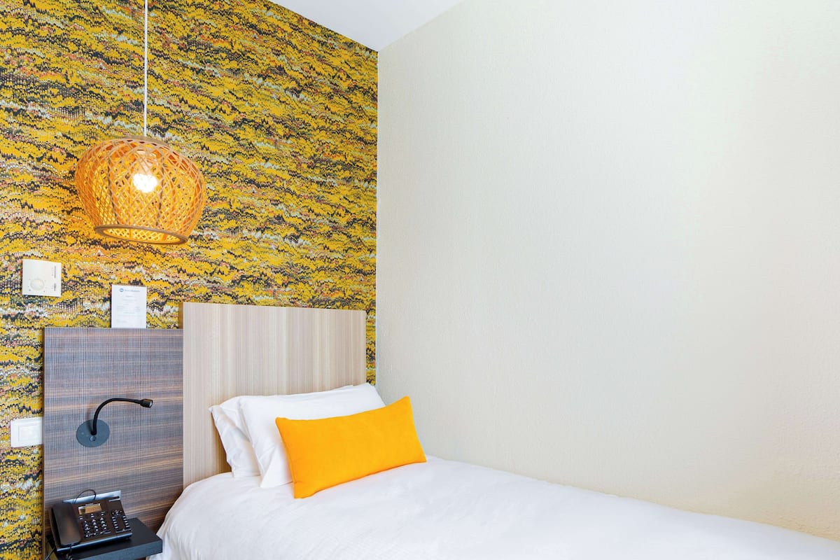 Habitación de hotel con papel tapiz estampado, cama con ropa de cama blanca y naranja, teléfono de noche y lámpara colgante.